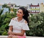 A cheerful woman sitting outdoors in a white lace top against a cityscape backdrop.